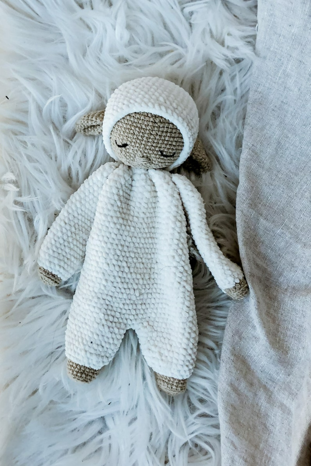 White textured crochet lamb with beige feet and ears asleep on a white fluffy blanket.