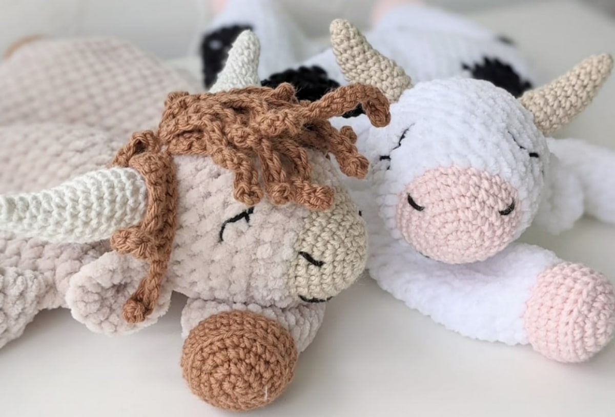 A black and white crochet cow with cream horns and its eyes shut next to a cream and brown cow with brown ringlets on its head.