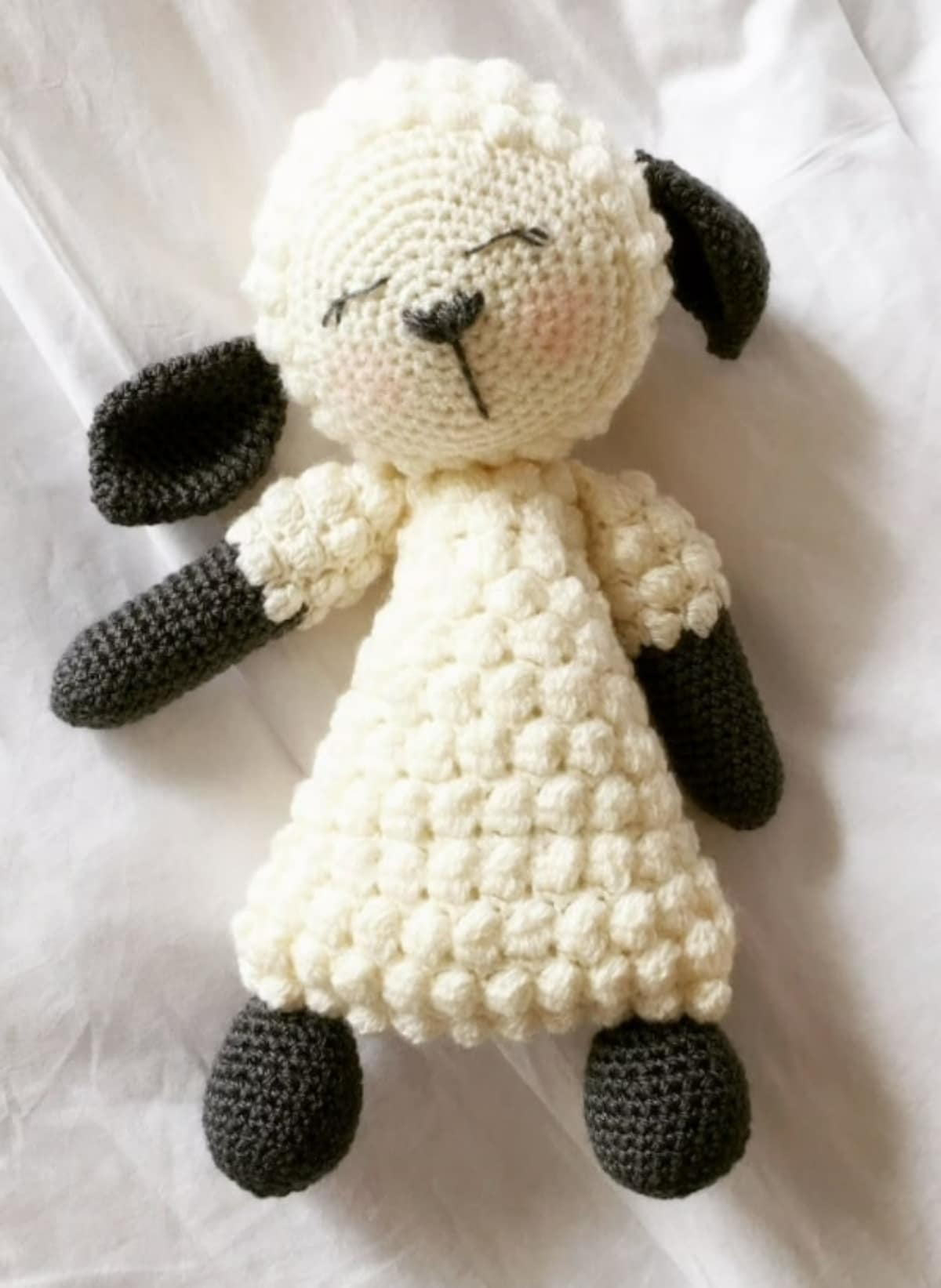 Small crochet lamb with white bobbles over its body and black ears and legs asleep on a white blanket.