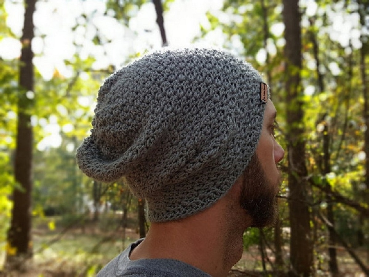 Brunette model side facing wearing a slouchy style gray crochet beanie and standing in a forest.