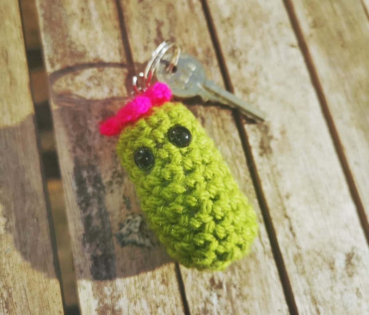 Small green crochet cactus with two eyes and a pink bow on its head attached to a keychain with a single silver key on a wooden table. 