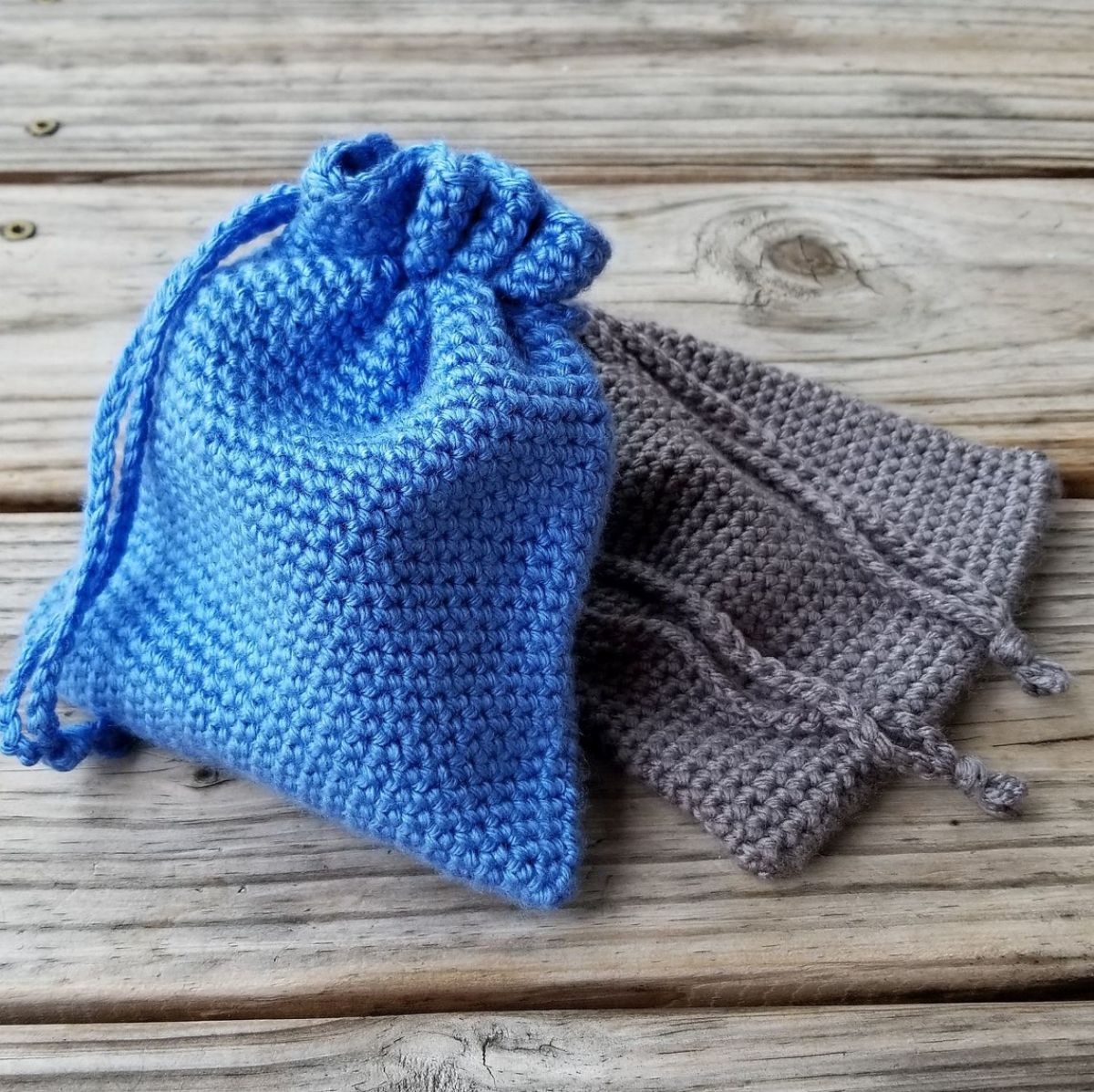  Small crochet blue drawstring bag sitting on a grey drawstring bag roughly the size of jewelry bags laying on a wooden table.