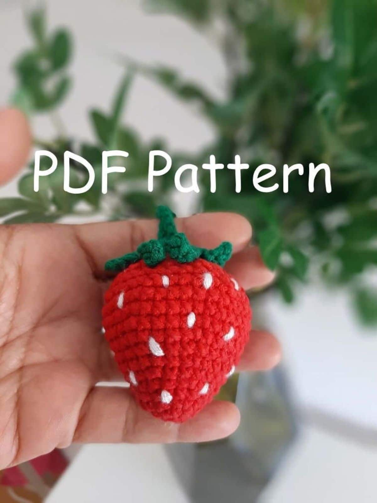 Small crochet strawberry with white seeds stitched in and a green stem laying on a hand with a white background with green foliage. 