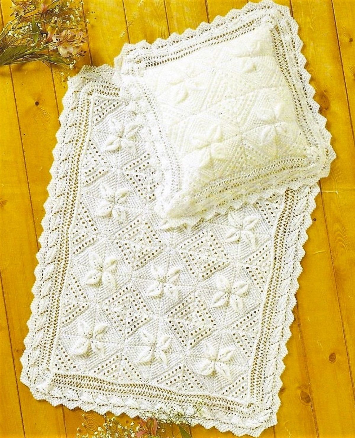 A white crochet lace baby blanket with a thick lace trim on all sides with a white cushion of the same design on top.