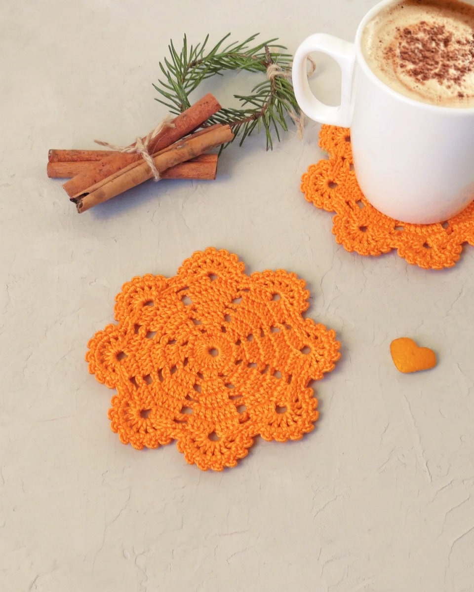 Two orange crochet snowflake shaped coasters, one with a white cup of cocoa on top next to some cinnamon sticks.