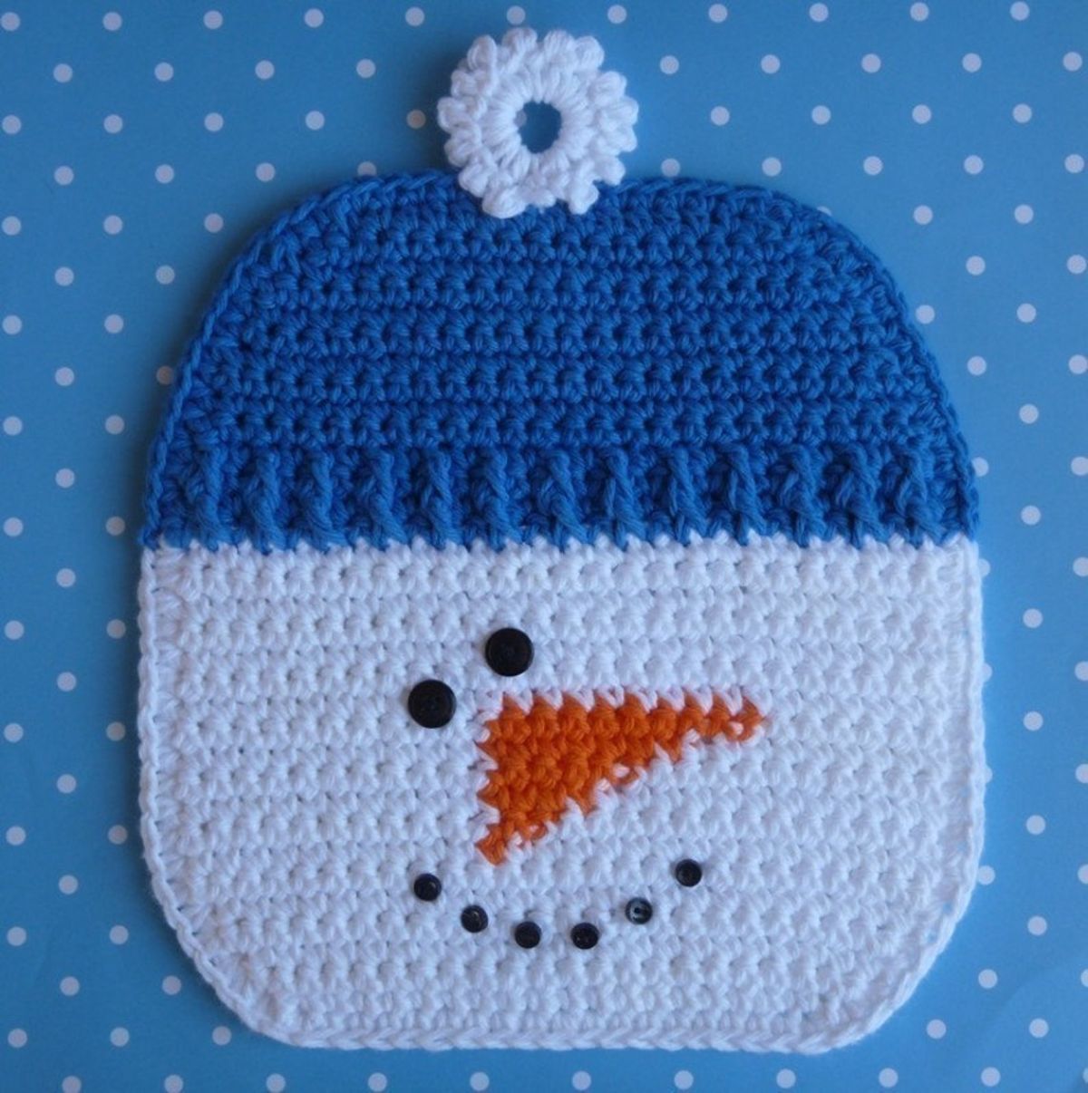 A white crochet snowman potholder with a blue beanie hat, orange carrot nose, and black eyes and mouth on a blue background.
