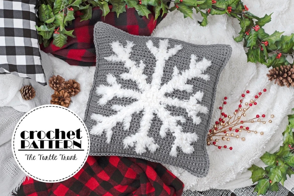A gray crochet cushion with a large white snowflake in the center on a wooden floor with holly around it.