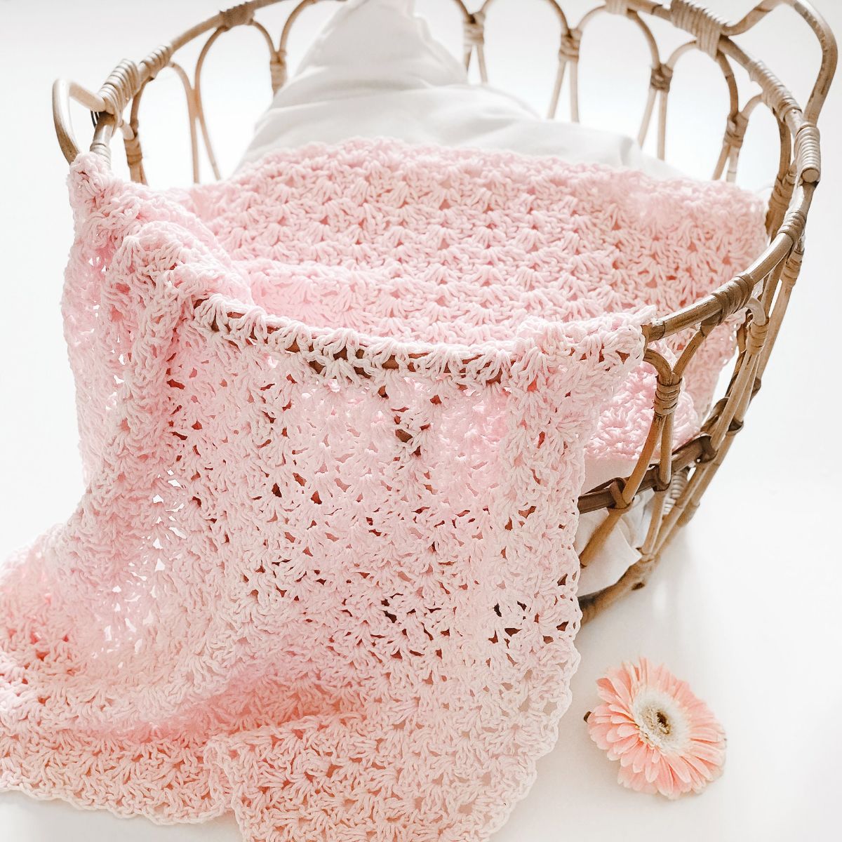 A brown wicker basket with a pink lacy crochet baby blanket spilling out of, next to a small pink flower.