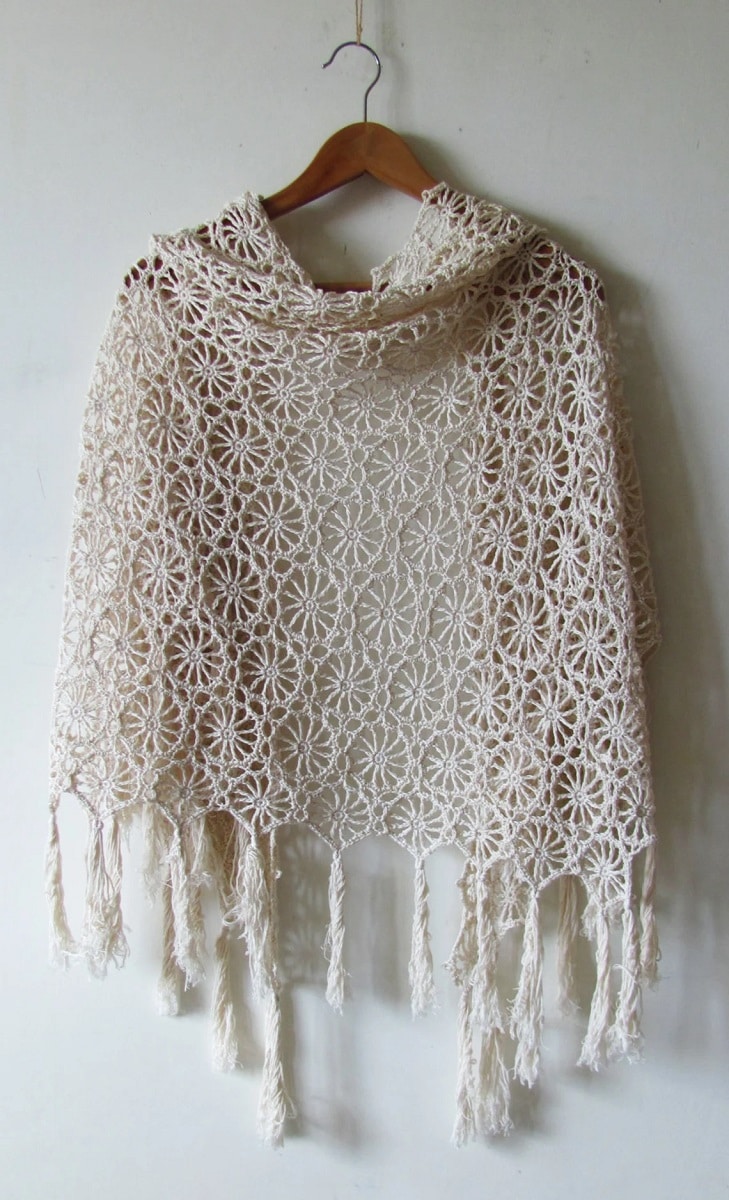 A cream crochet shawl with a circular flower pattern all over and long thick tassels at the bottom on a wooden hanger.