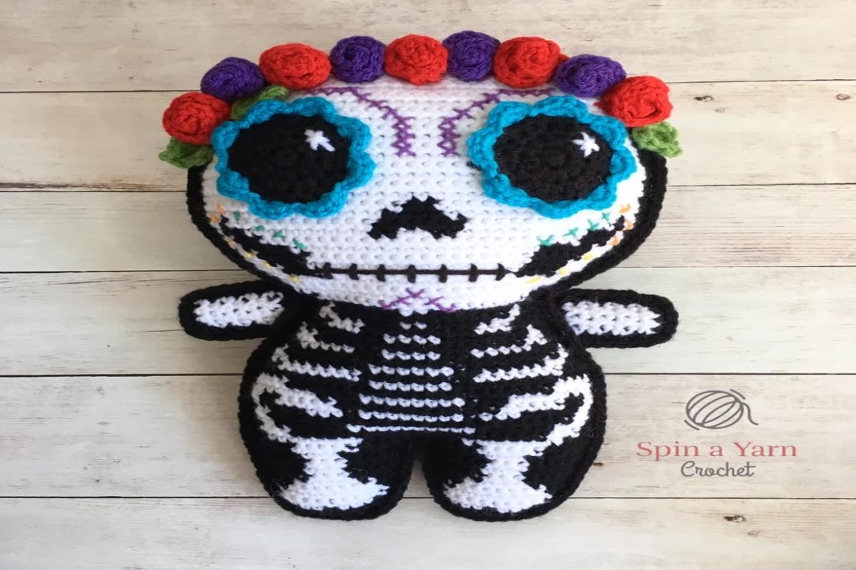 Small crochet skeleton doll with an oversized skull, blue flowers for eyes, and small pink and purple flowers along the top of the skull.