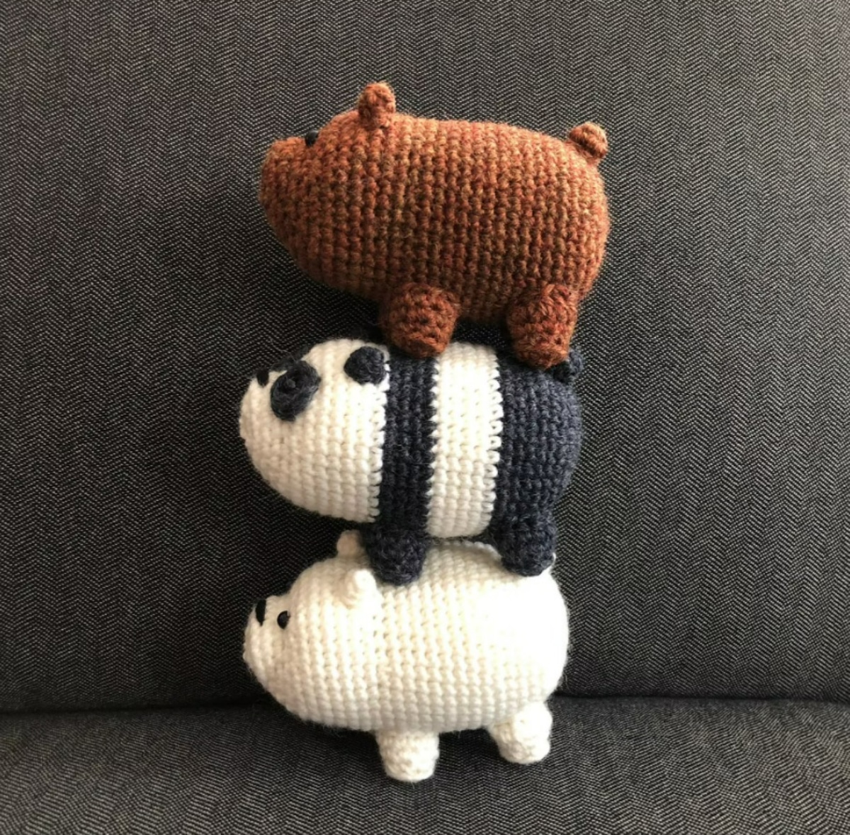 Three crochet bears stacked on top of eachother. A white bear, black and white bear, and brown bear on a gray sofa.