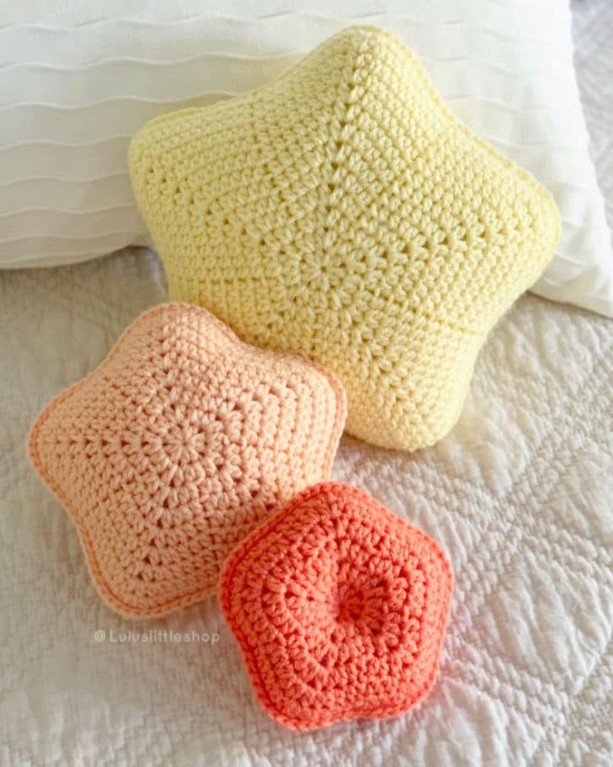 A small coral star shaped crochet cushion in front of a peach and larger yellow cushion. All cushions are stuffed and sit on white bedding. 