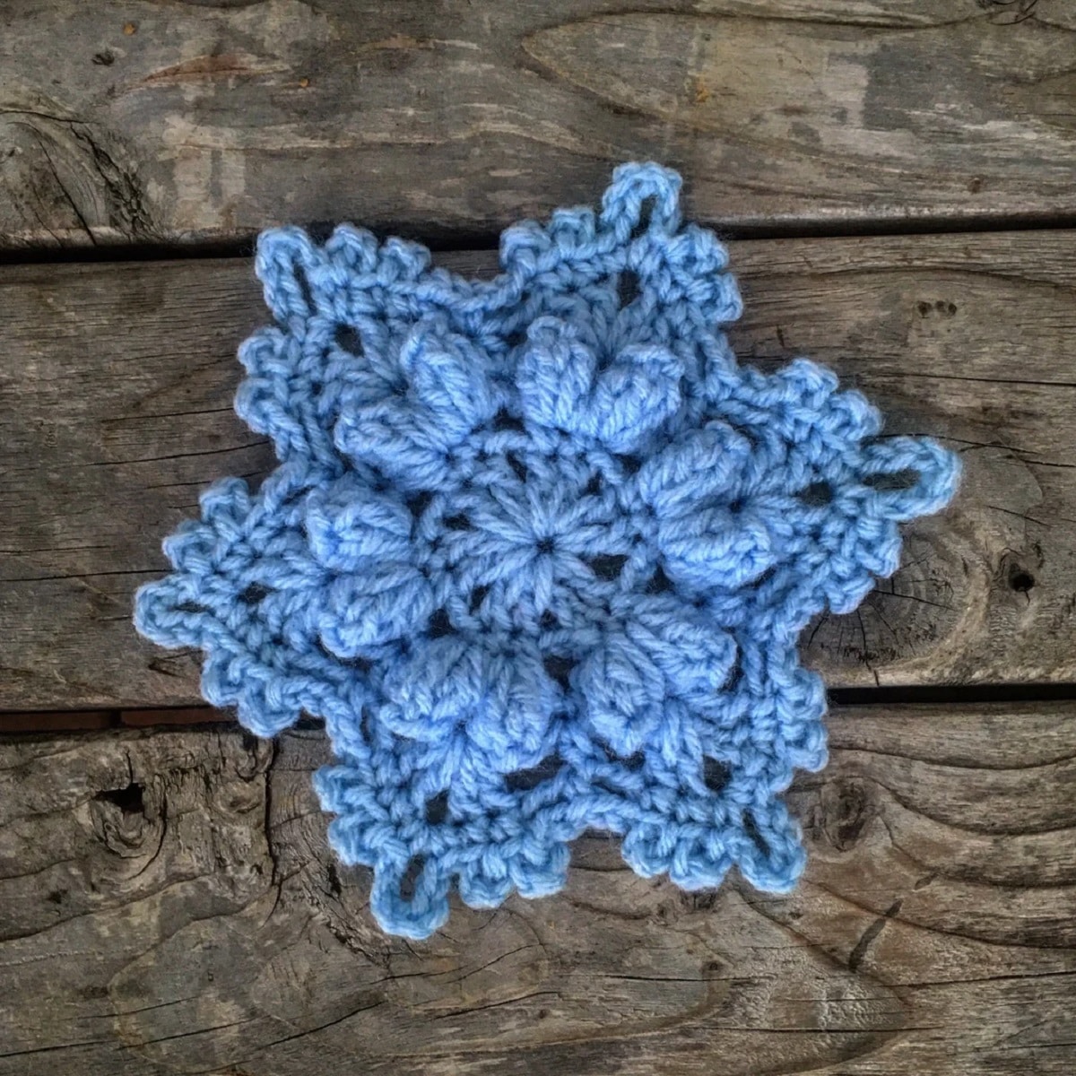 Blue crochet snowflake with a small face trim on each side on a wooden floor.