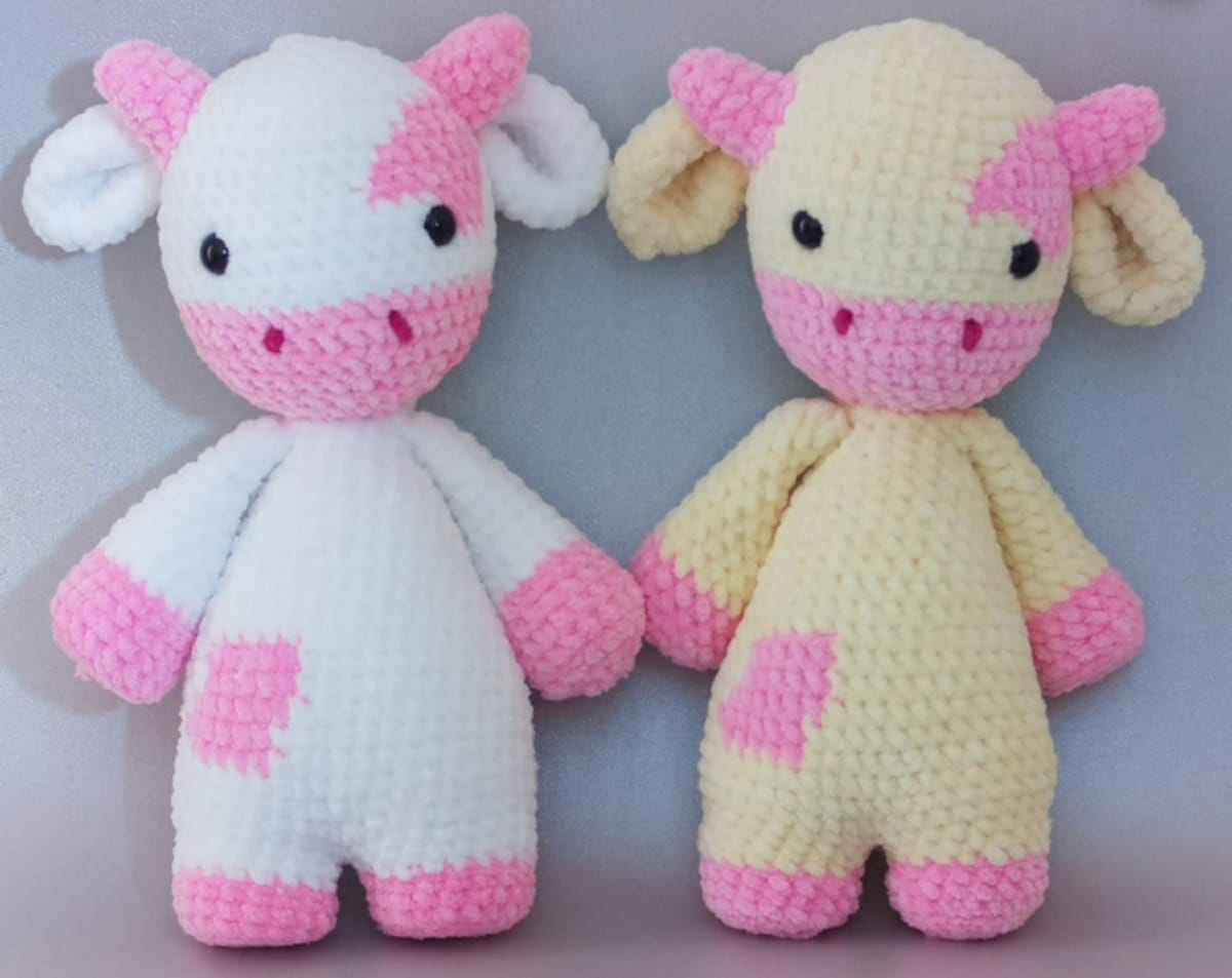 A large white crochet cow standing on two legs with pink horns, hands, feet, and a patch on its side next to a yellow and pink cow of the same design.