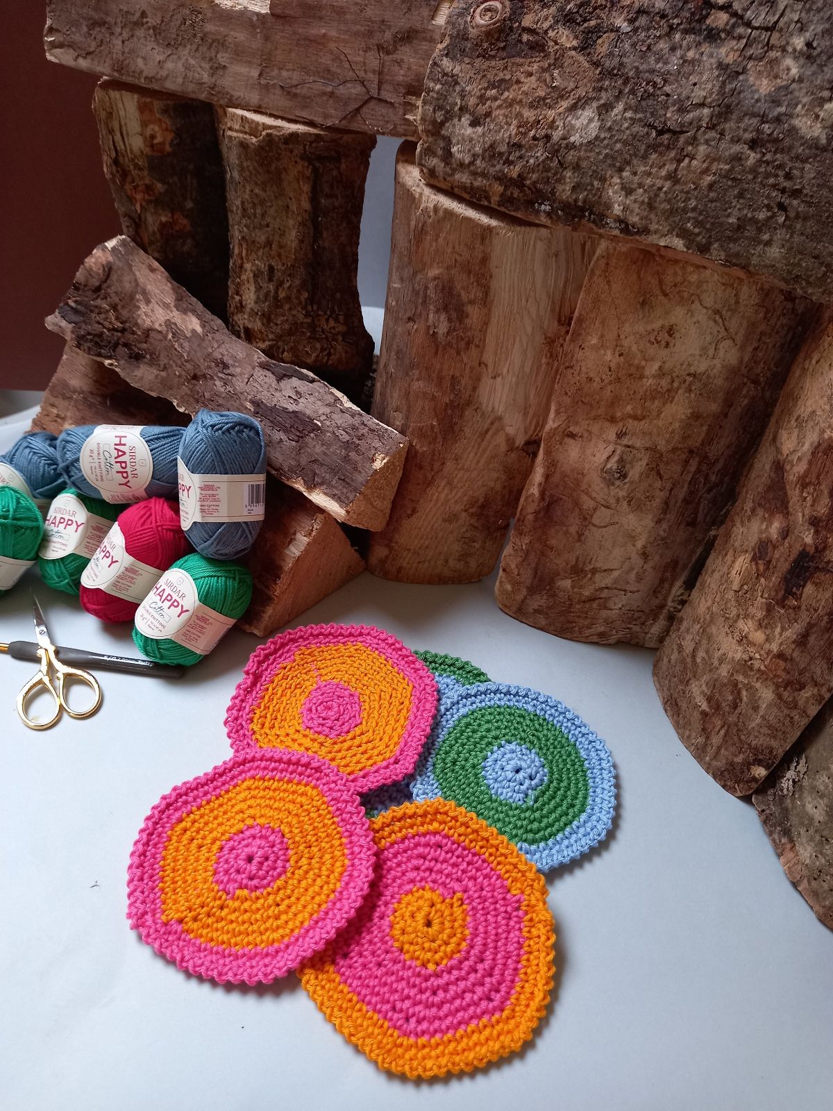 Orange and pink small round crochet coasters on top of blue and green coasters next to firewood and a small pile of pink, blue, and green yarn. 