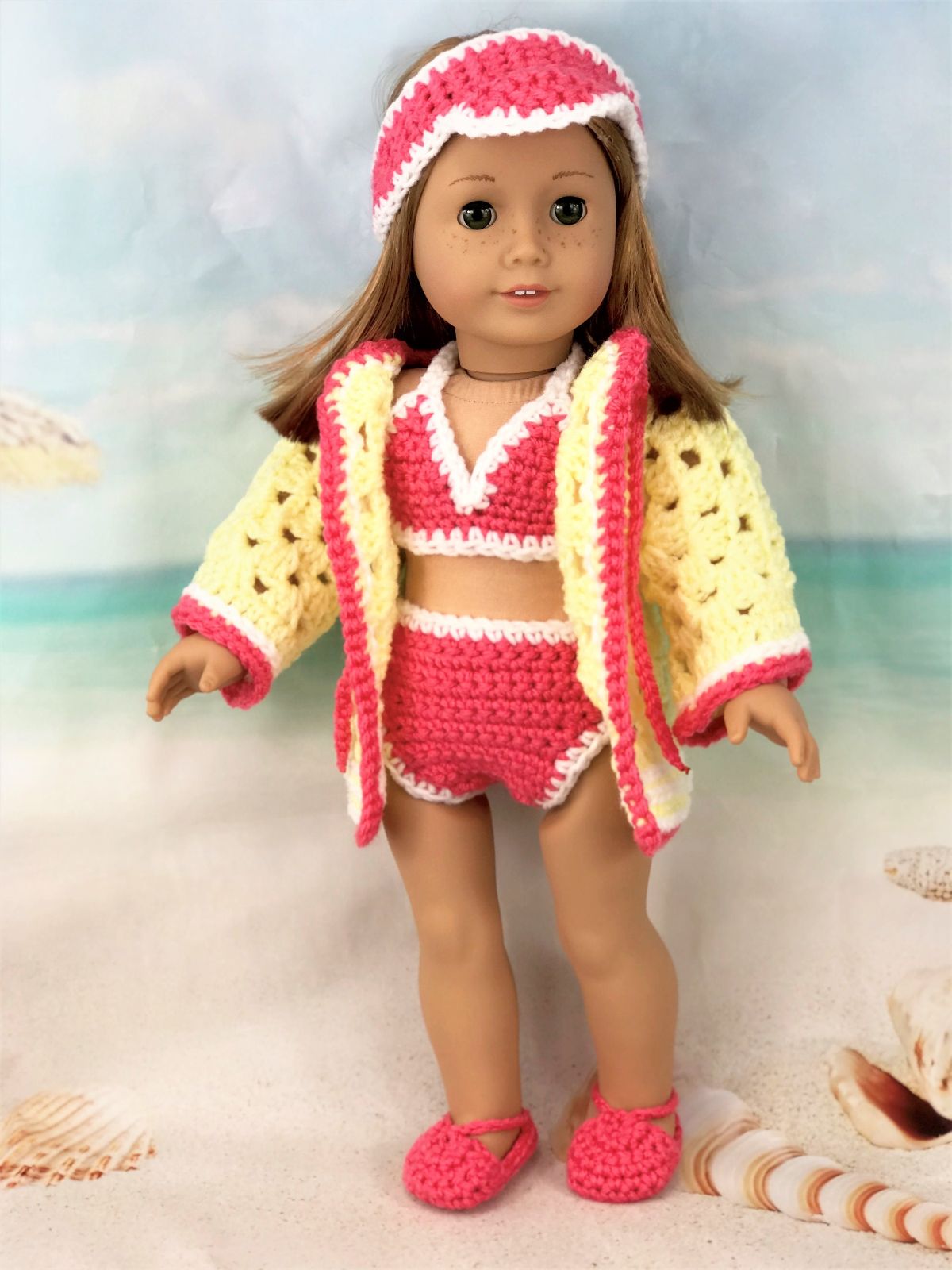 An American doll wearing a pink and white crochet headband, matching bikini set, and a yellow and pink cardigan with pink slip-on shoes.
