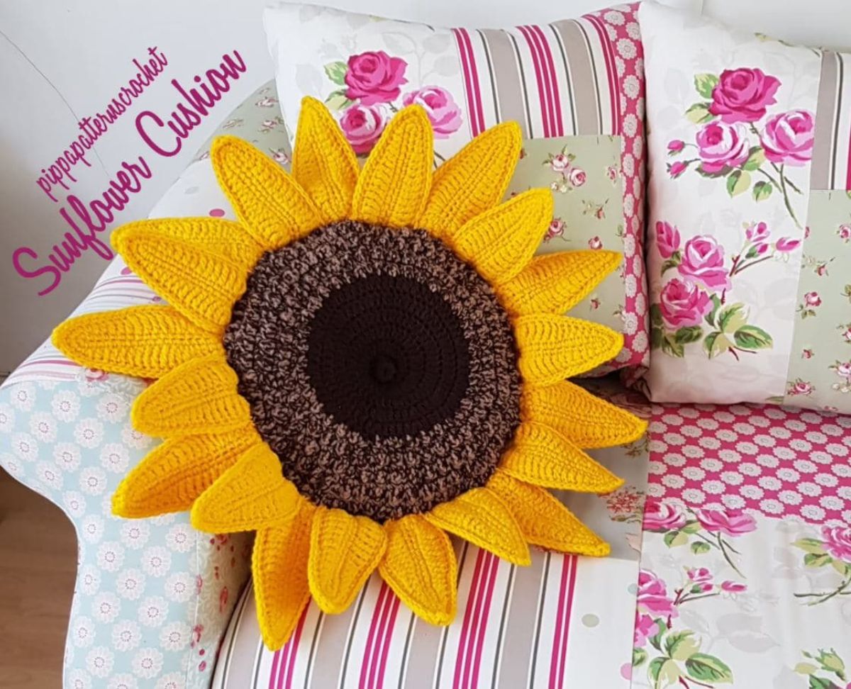 Large crochet sunflower shaped cushion with yellow petals and a brown center sitting on pink floral and striped cushions. 