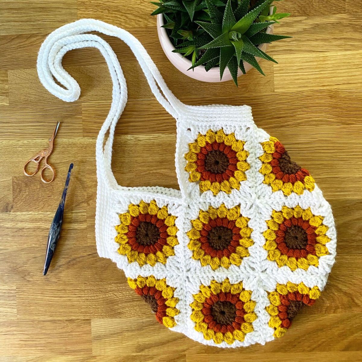  White granny square style crochet bag with sunflower design and long white handles on a wooden floor. 