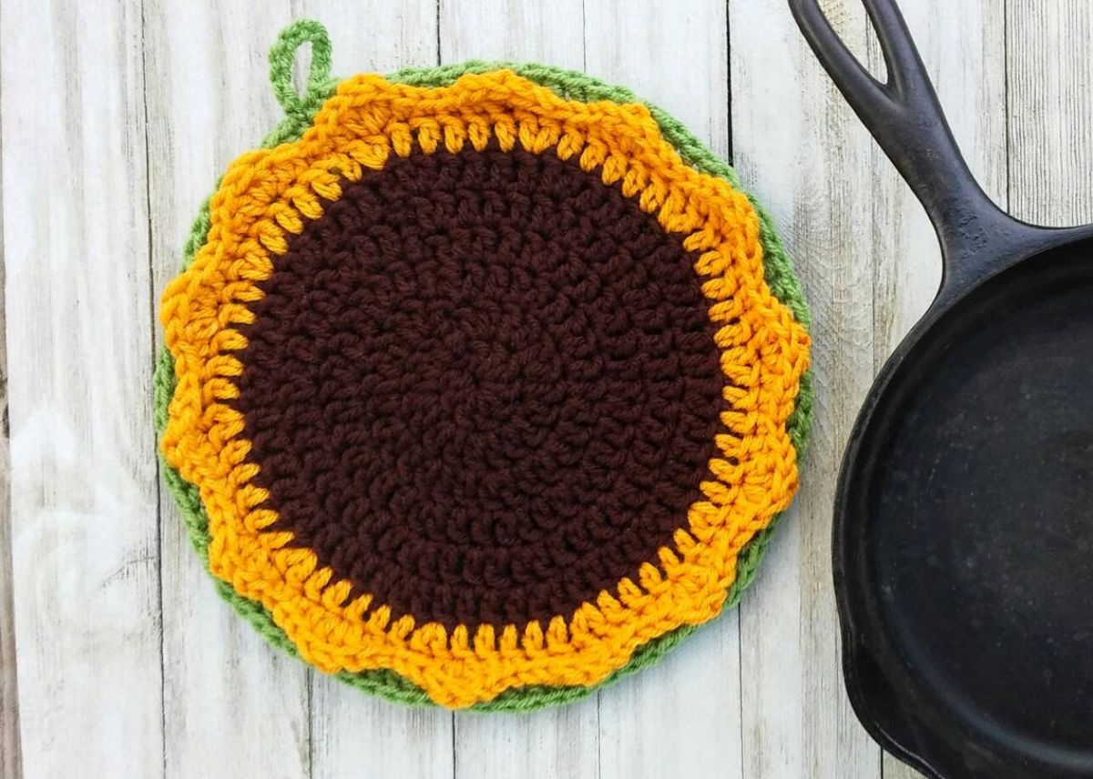 Large sunflower shaped crochet pot holder with a small green loop at the top next to a black frying pan.