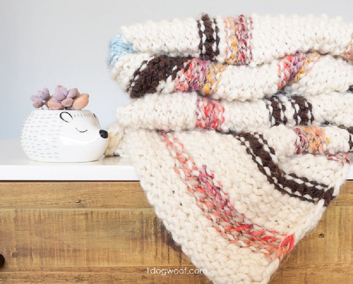 Cream crochet blanket with pink and black horizontal stripes across it folded on a cupboard.