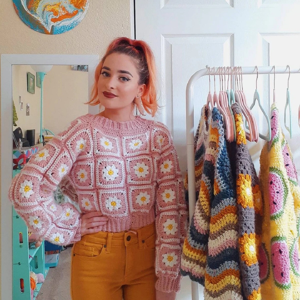 Redhead woman wearing a pink crochet sweater with white and yellow daisies in squares all over the sweater.