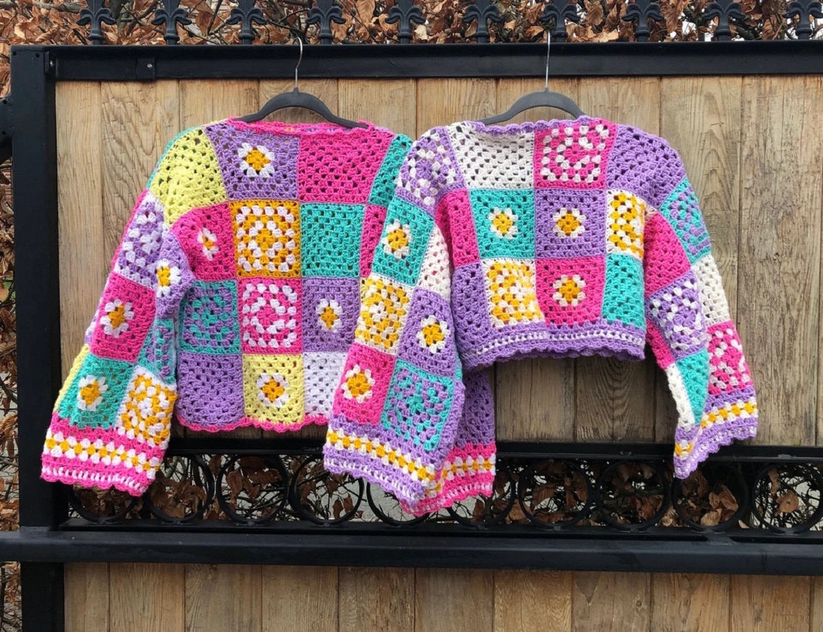 Two patchwork pastel colored crochet sweaters with small flowers in the squares on hangers by a wooden fence.