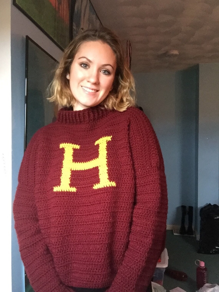Blonde woman wearing a maroon crochet sweater with a large yellow ‘H’ stitched across the chest.
