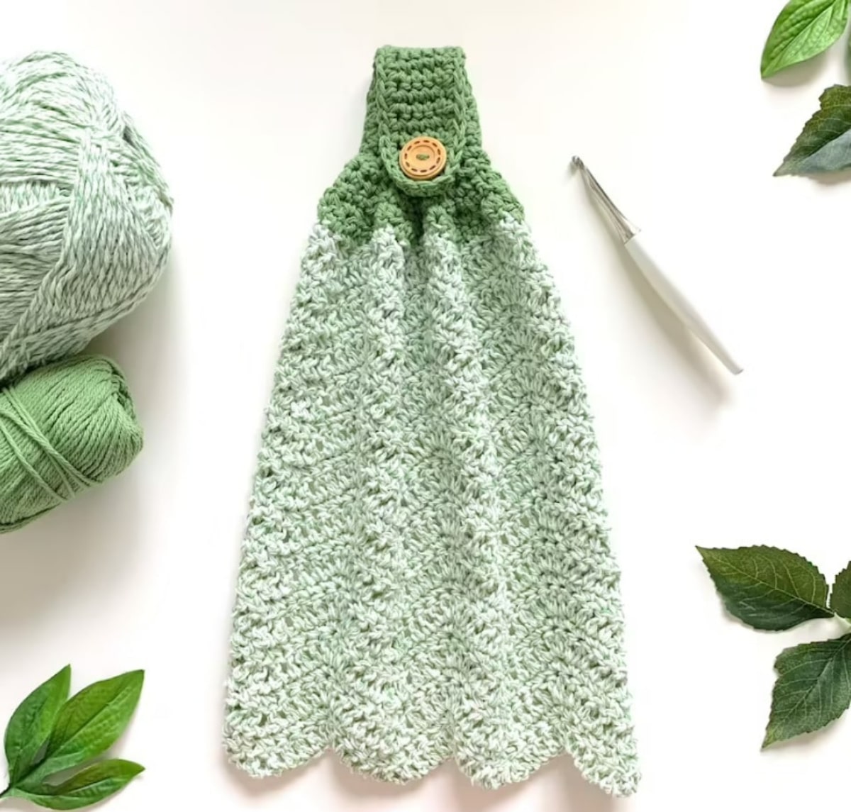 A light green crochet hand towel with a scalloped edging and a dark green loop with a button at the top.
