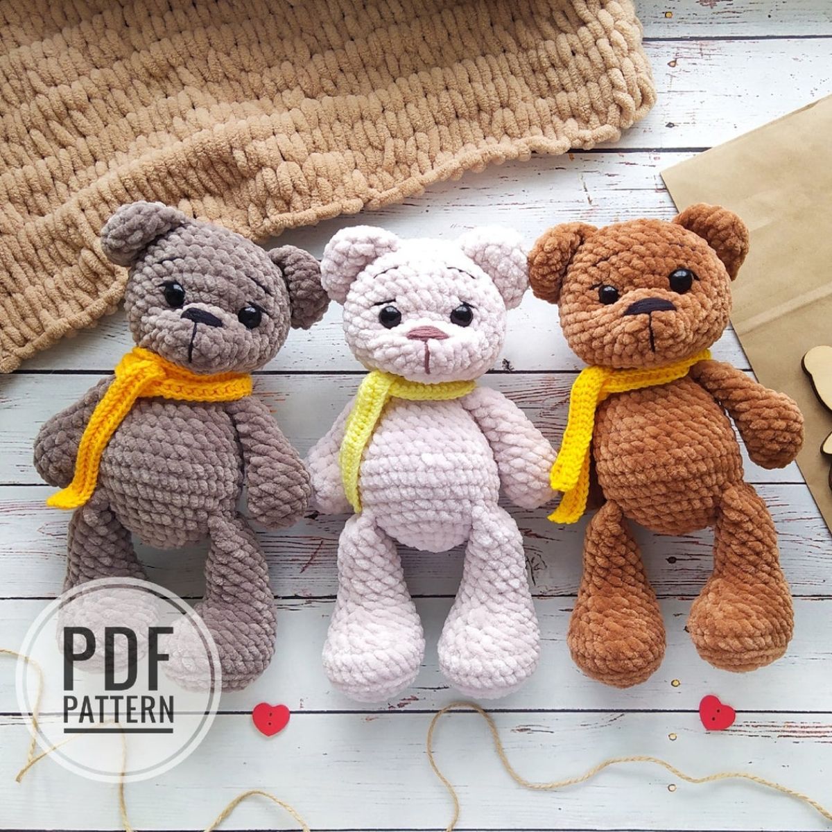 A light brown, white, and dark brown crochet teddy bears all wearing yellow scarves lying in a line on a white wooden floor.