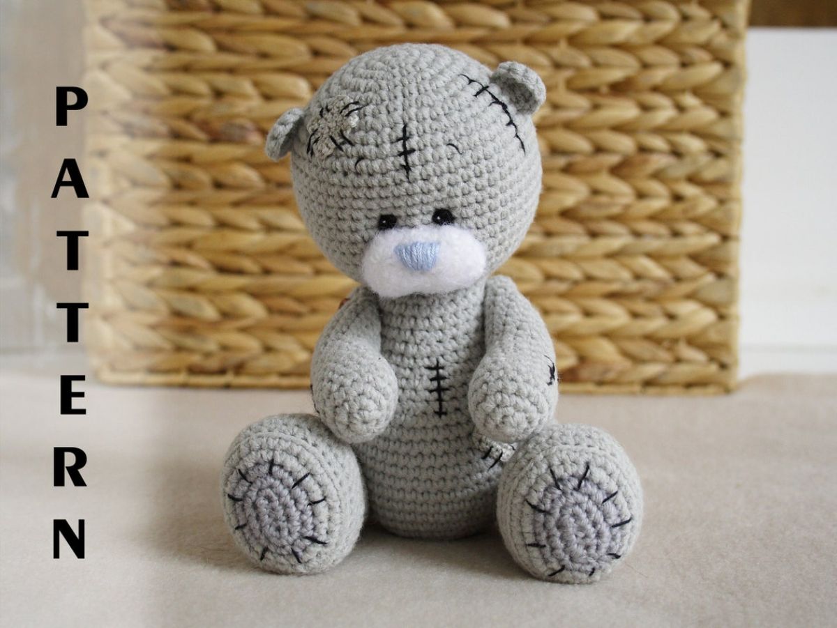 A pale green crochet teddy bear with a small blue nose and black stitches on its head, body, and feet sitting down.