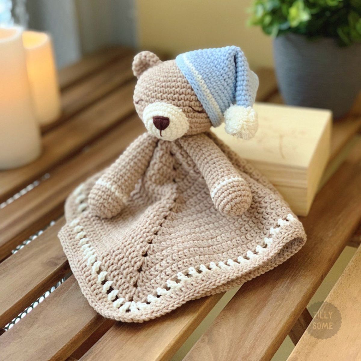 Small brown crochet blanket with a teddy bear's face and arms attached, wearing a blue night cap over its right ear.