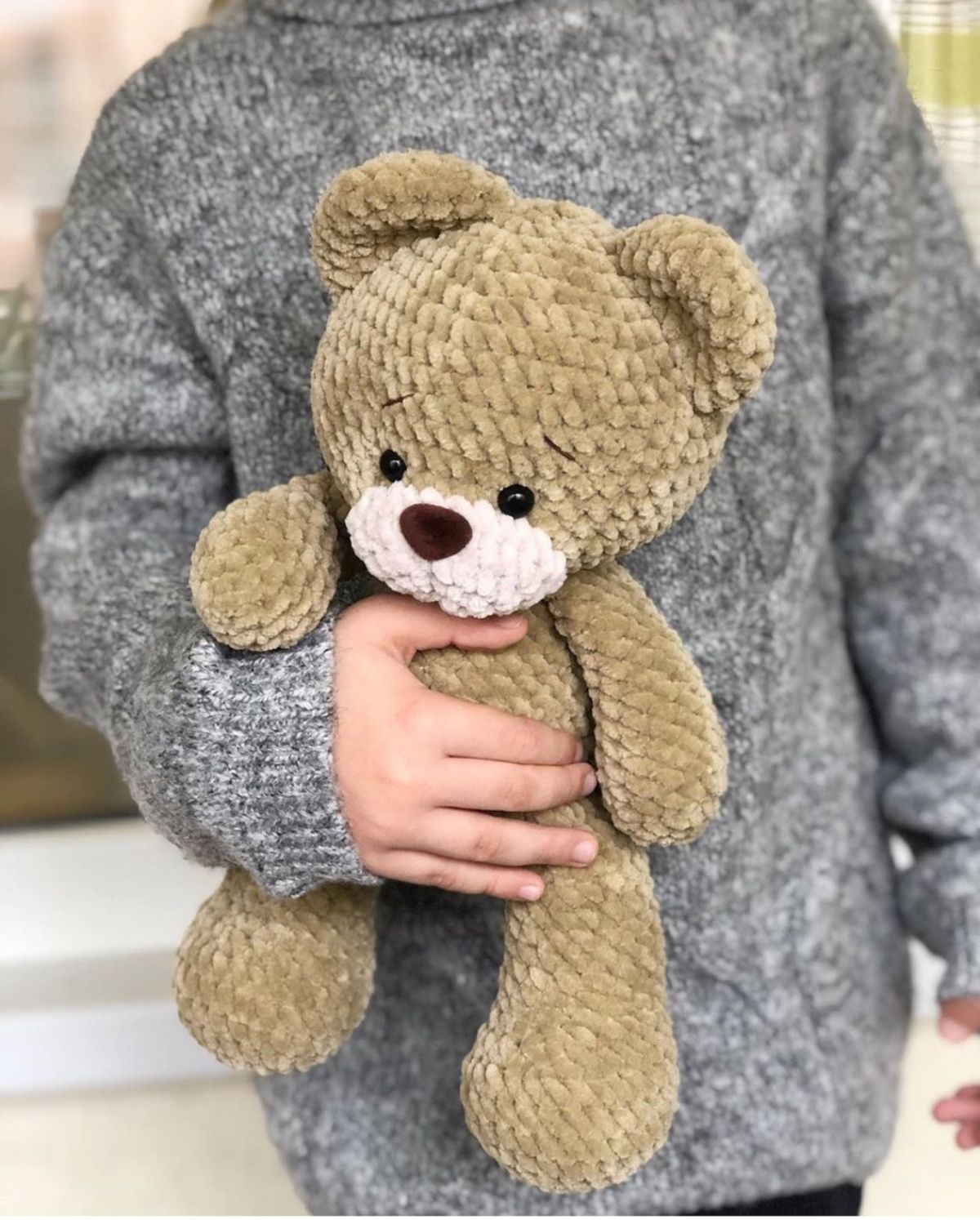A hand holding a large crochet teddy bear with a dark brown nose, white mouth, and a light brown face, body, and legs.