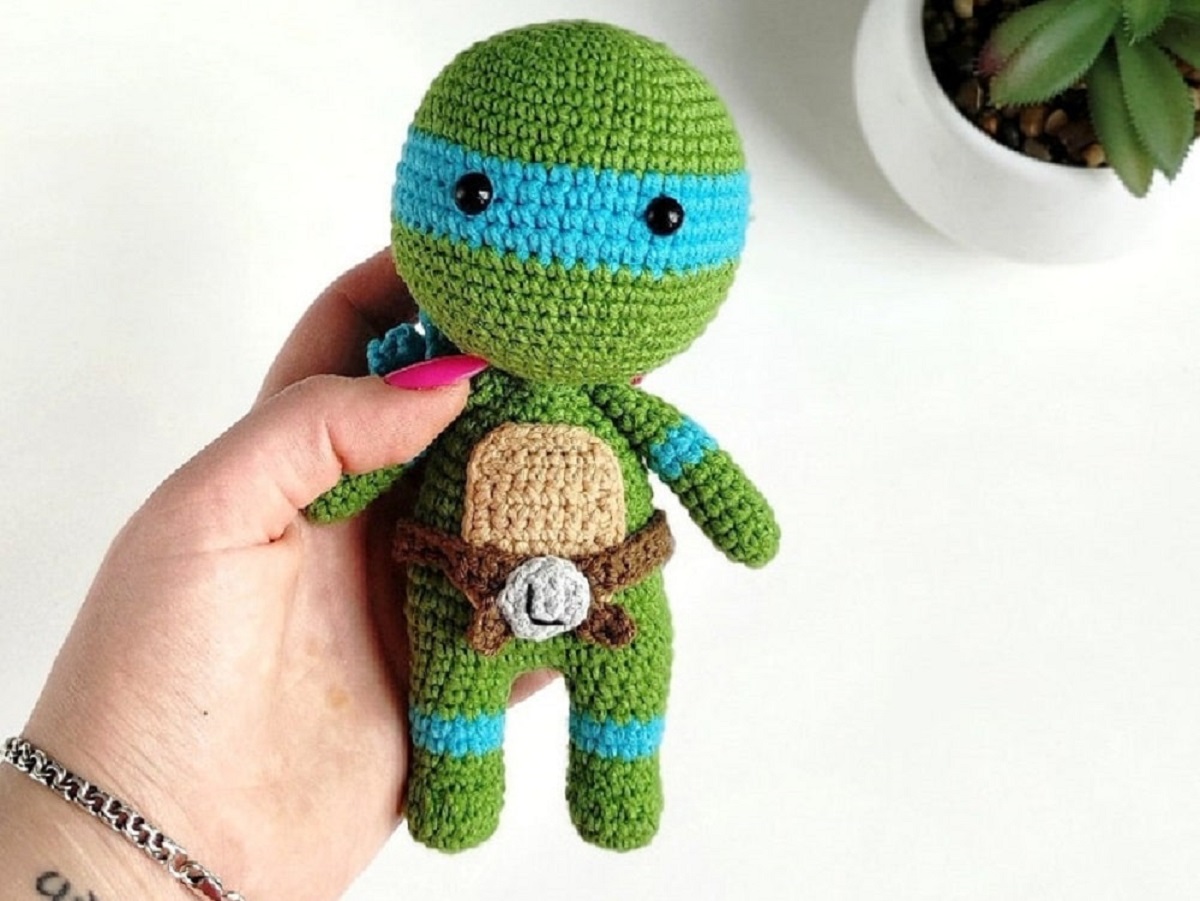Blue, green, and brown crochet stuffed teenage mutant ninja turtle wearing a brown belt and held in a manicured hand. 