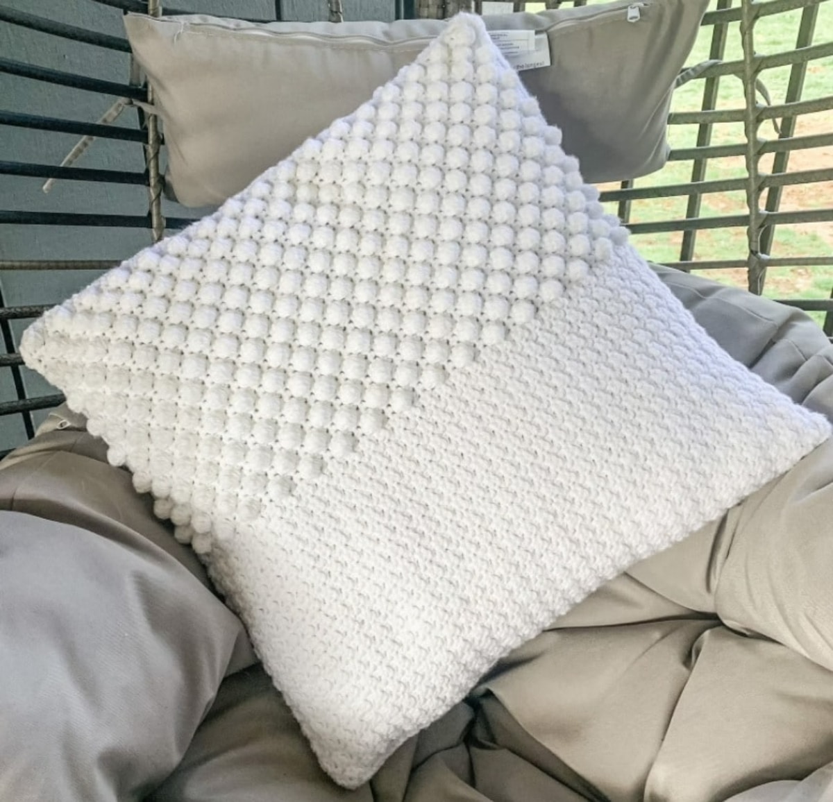 Cream crochet cushion with one half covered in textured bobbles and the second half is plain placed on a gray chair. 