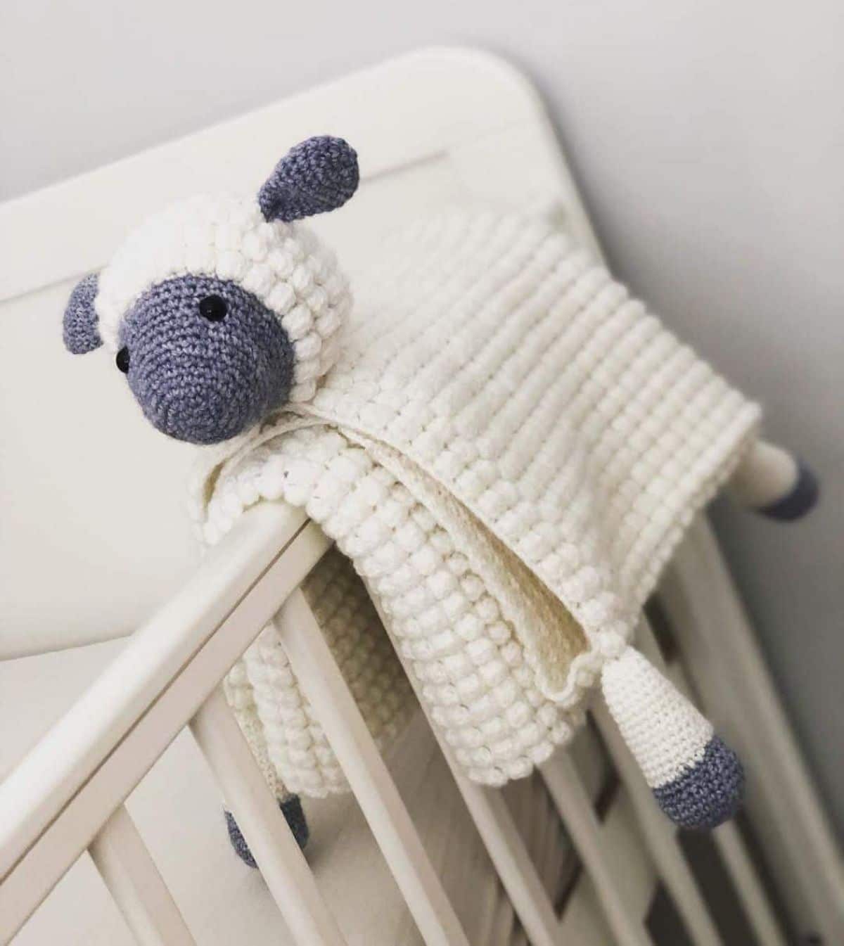 Cream crochet blanket with stuffed sheeps head and legs attached folded and draped over the railings of a cream crib. 