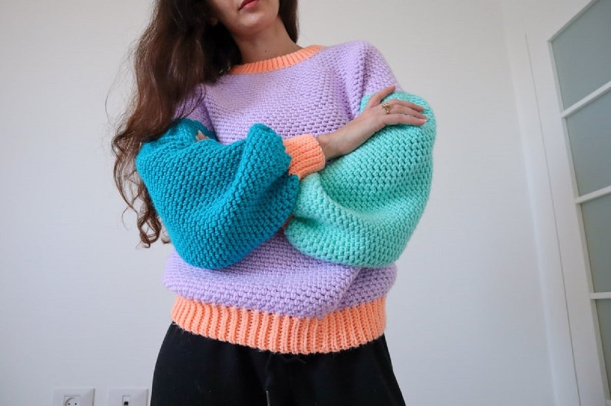 Brunette woman wearing an oversized lilac crochet jumper with an orange collar and hem and dark and light blue long sleeves.