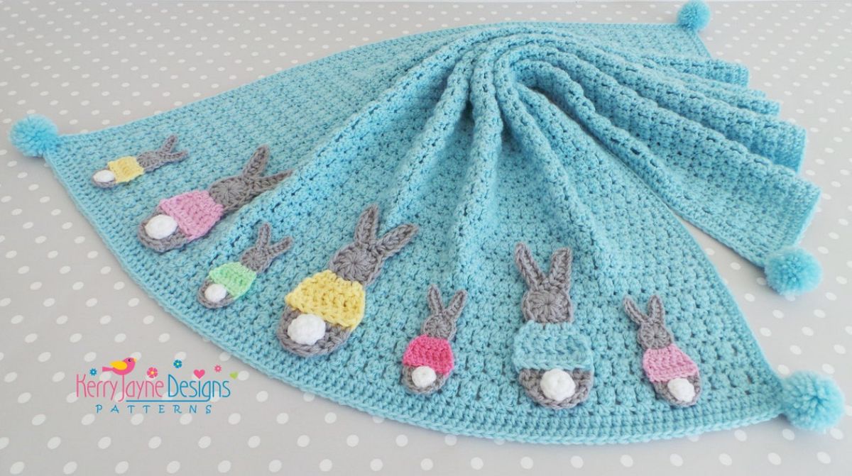 Blue crochet baby background with adult and child-sized bunnies wearing different colored jumpers with white tails across the bottom. 