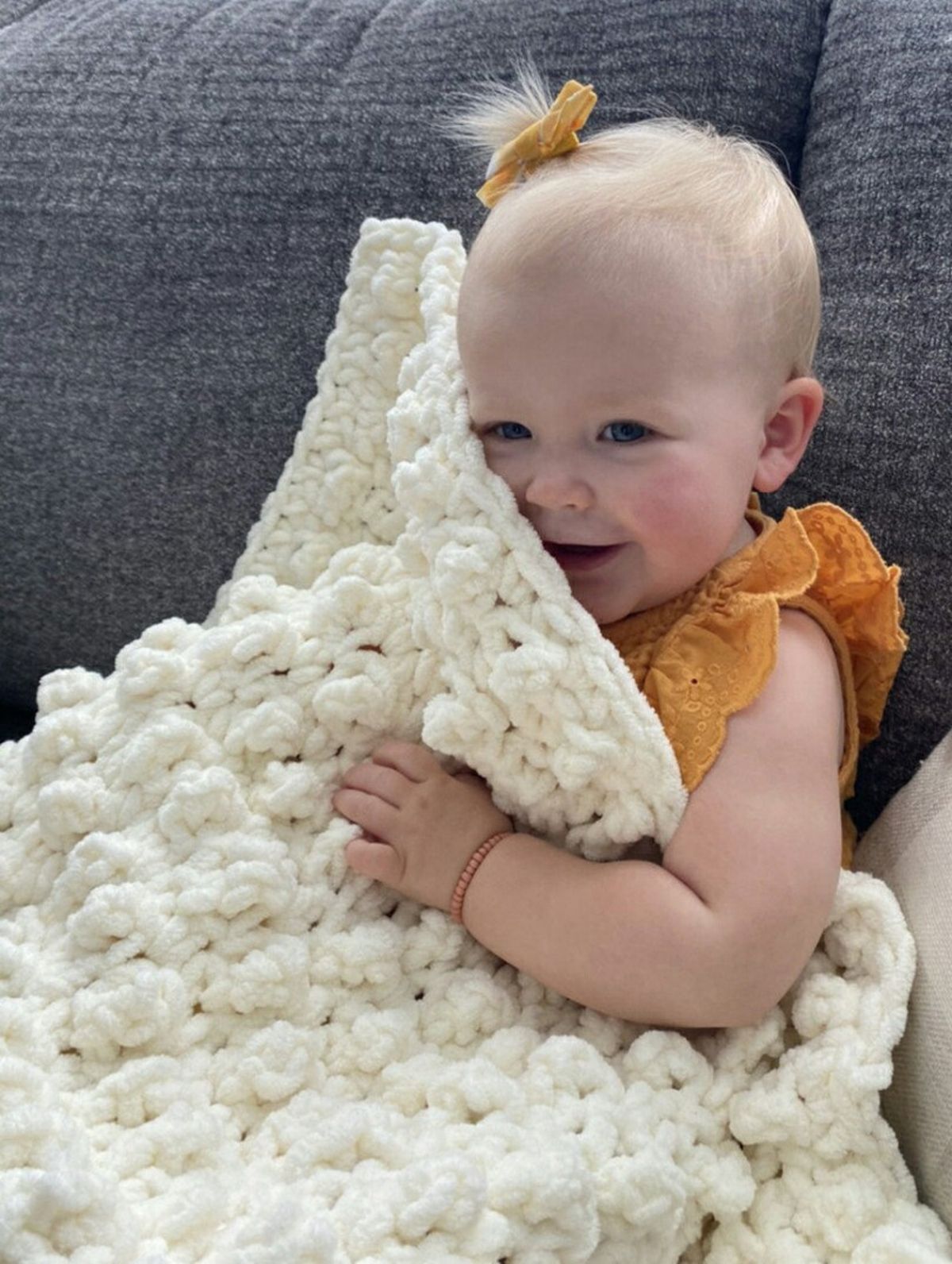 Smiling baby in a yellow dress and matching scrunchie under a chunky cream crochet blanket sitting on a gray fabric sofa.