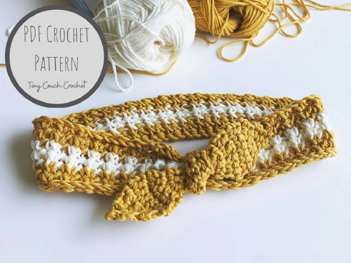 A thin white crochet headband with a white band through the middle and a small yellow bow in the front laid on a white background with yarn balls behind it. 