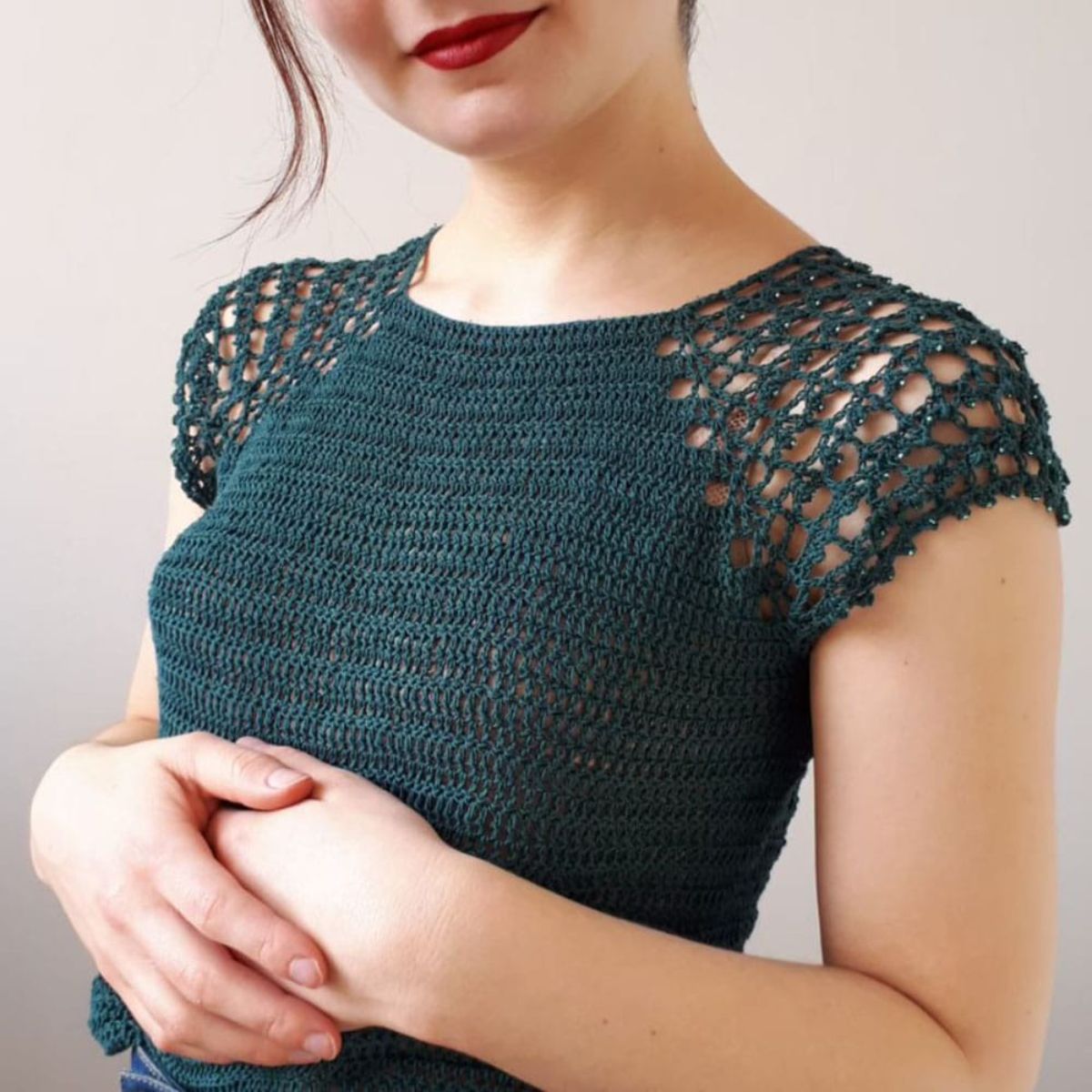 Brunette woman in red lipstick and arms across her stomach wears a dark green crochet top with short wide knit sleeves. 