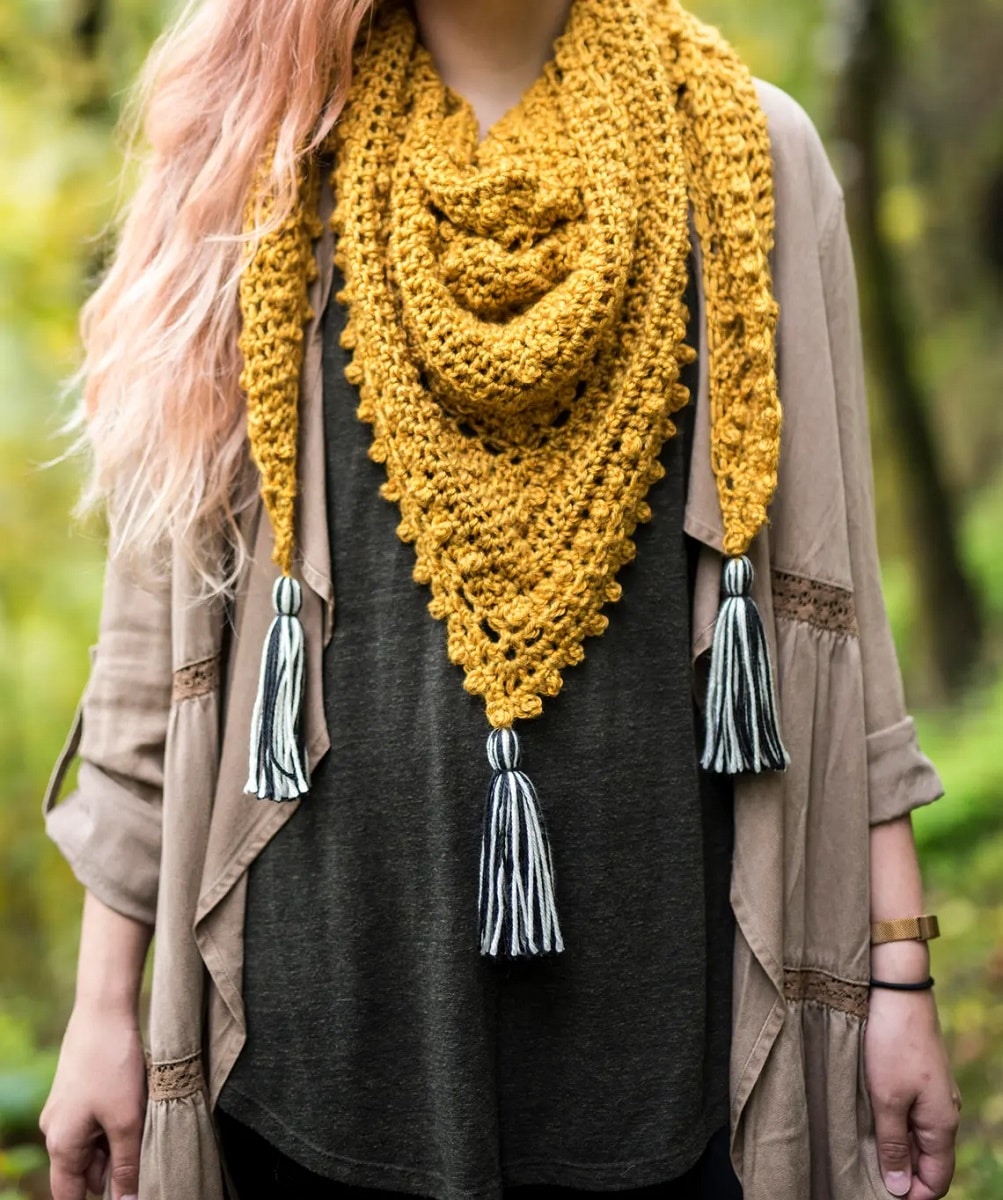 Blonde woman wearing a mustard yellow crochet triangle scarf with bobbles on the edges and three blue tassels hanging from each corner.