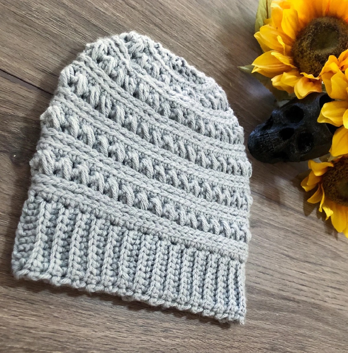 A light green crochet beanie hat with a thick banding around the bottom next to some sunflowers on a wooden table.