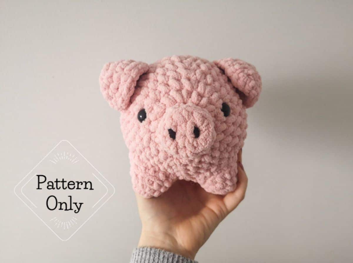 Pale pink crochet and stuffed pig with black nostrils and eyes held up and displayed with a white background.
