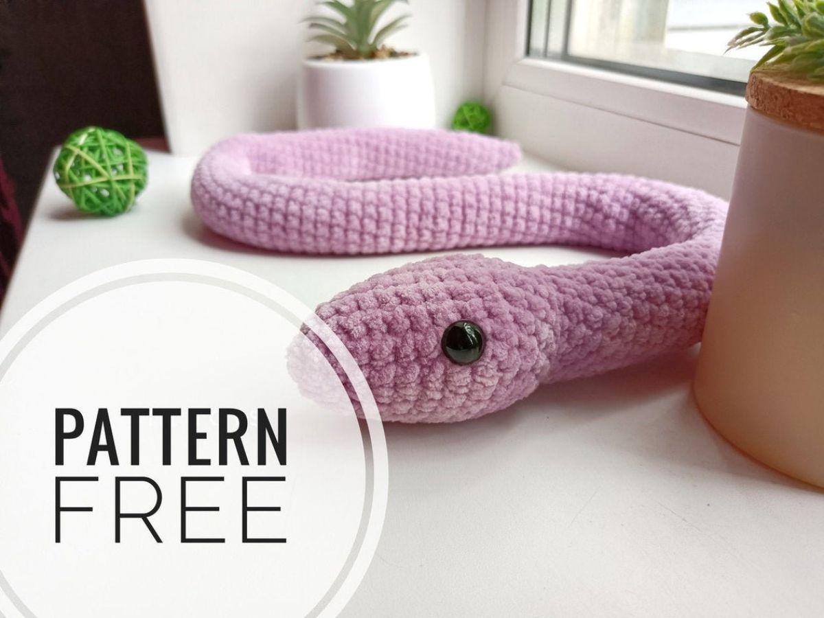Pink crochet stuffed snake plush with black eyes attached curled up on a window sill next to some potted plants. 
