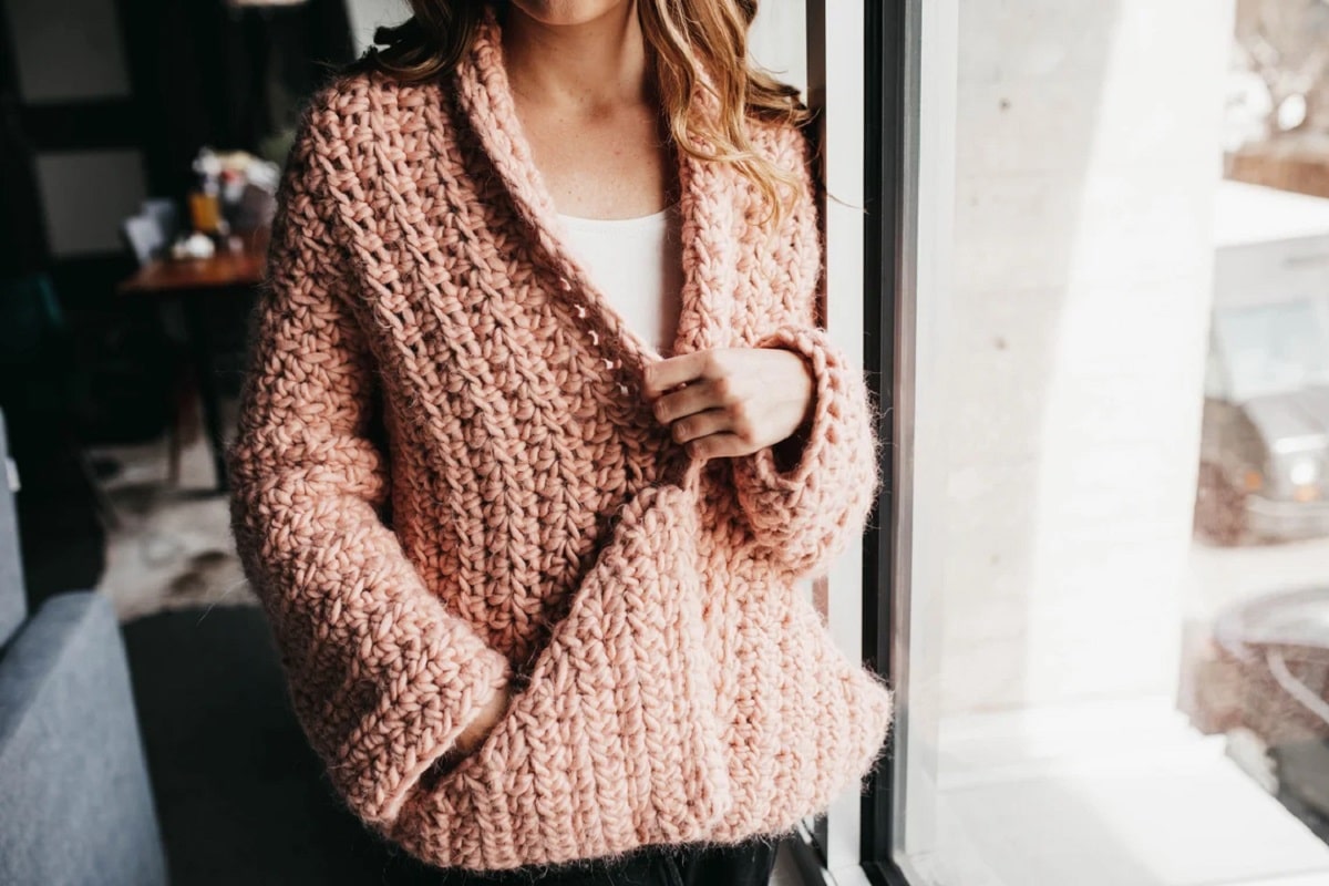 Slim woman wearing a chunky salmon crochet cardigan with one hand in a large pocket standing next to a large window. 