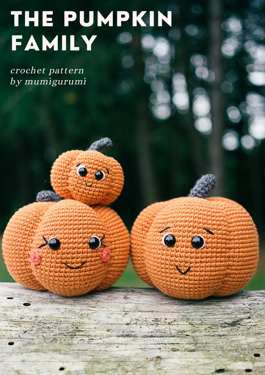 Two large crochet orange pumpkins with smiling faces sitting on a log with a smaller pumpkin on top of one pumpkin.