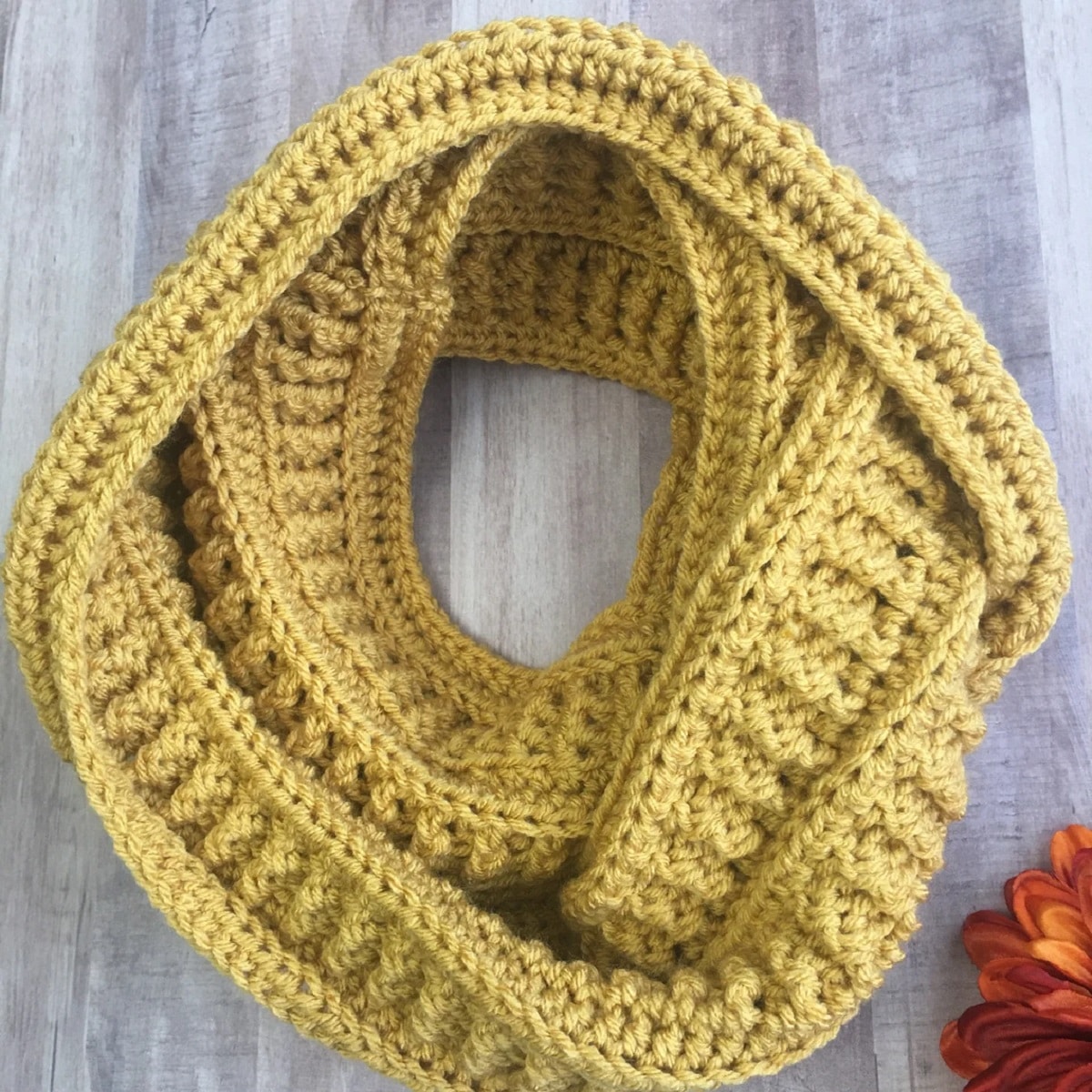  Yellow crochet infinity scarf with thick vertical lines and a horizontal trim wrapped into a circle on gray wooden flooring.