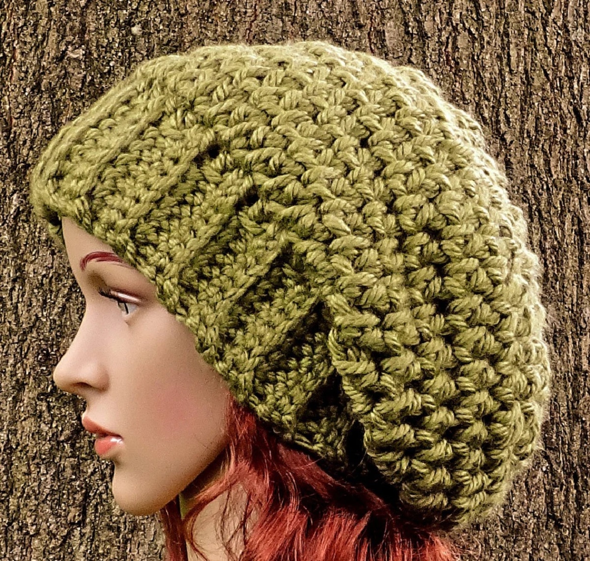 Slouchy olive green crochet hat worn by a mannequin with red hair in front of a tree. 