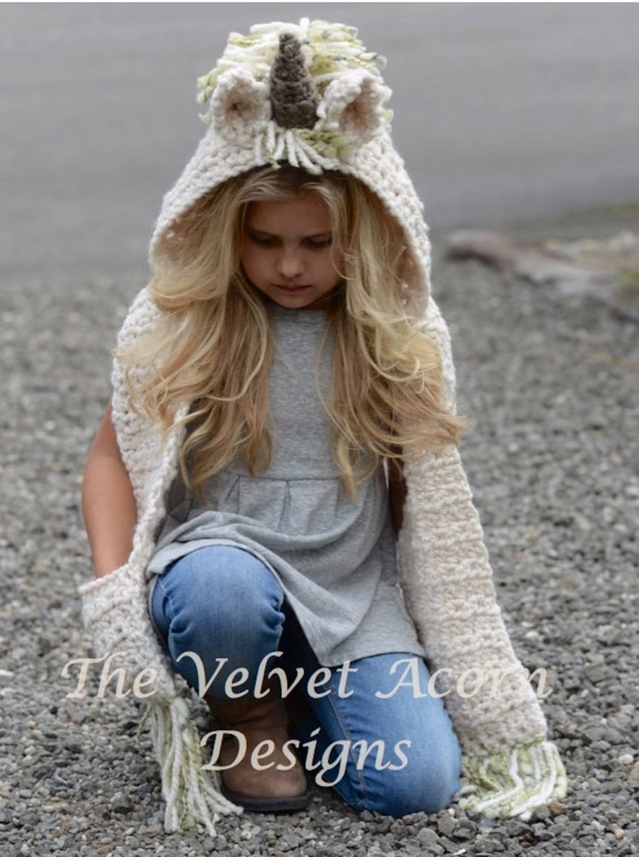 Young blond girl wearing a cream crochet unicorn hood with horn, ears, and mane with pockets attached kneeling on small gray stones. 