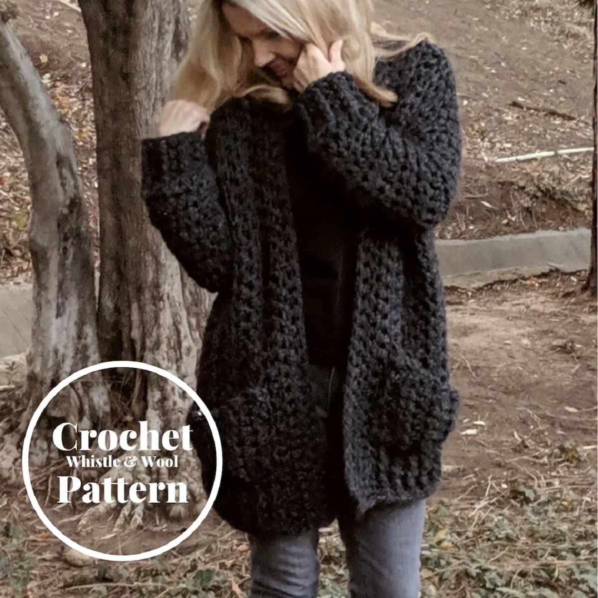 Large black crochet cardigan with long sleeves and pockets worn by a blond woman standing in front of a tree. 